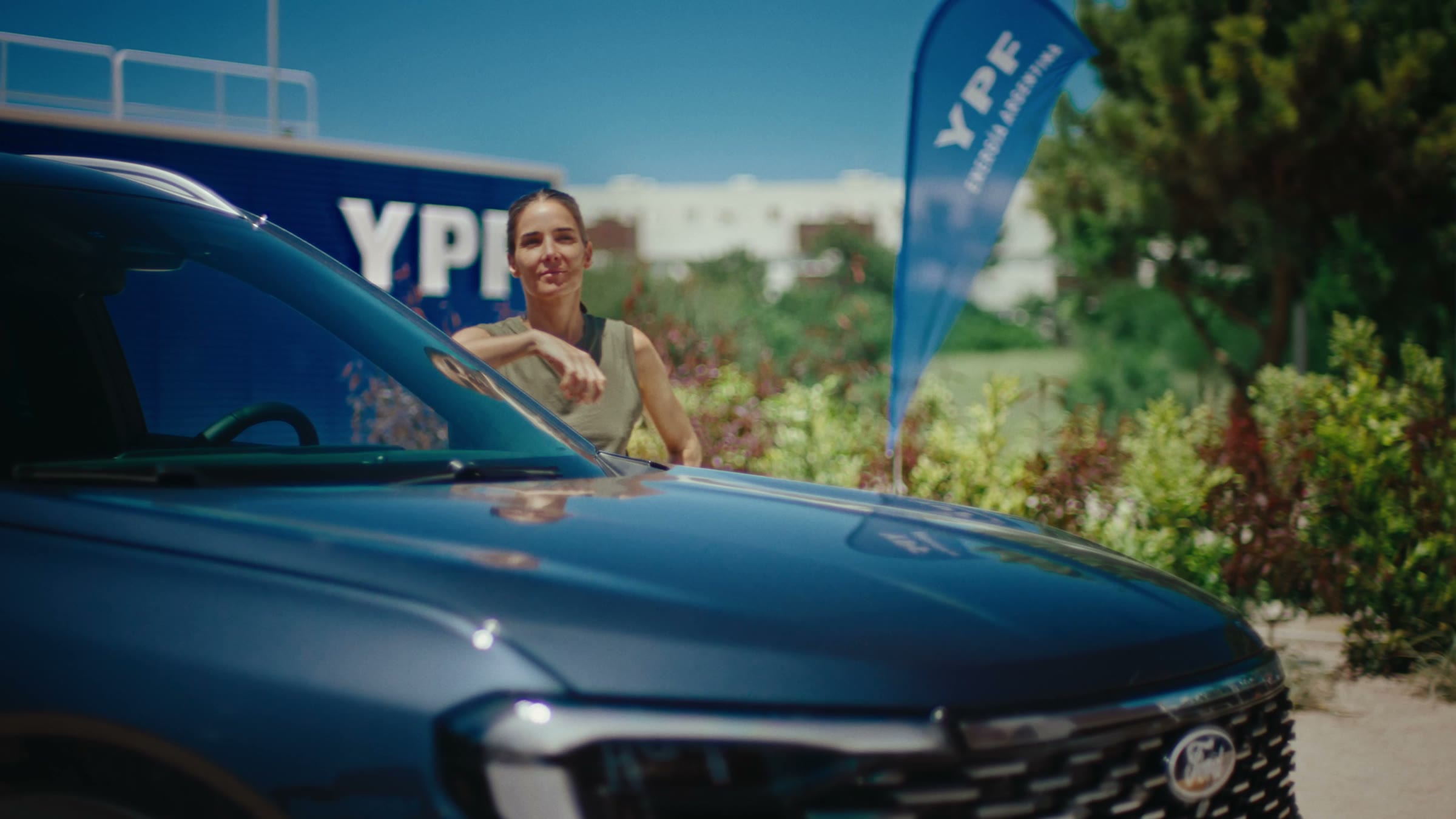 Juana Viale leaning on a car hood in summer light, YPF station behind